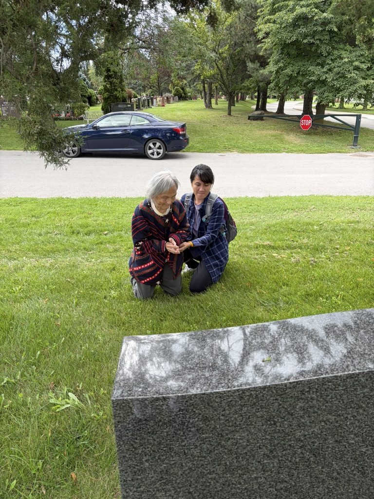 Helen kneeling on the grass beside David's headstone at Pine Hills Cemetery, with a family member crouched beside her.