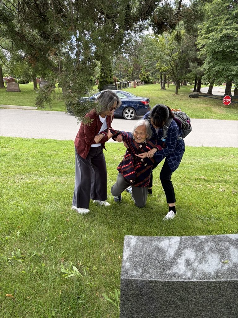 Helen laughing as two family members help her up from the grass beside the headstone.